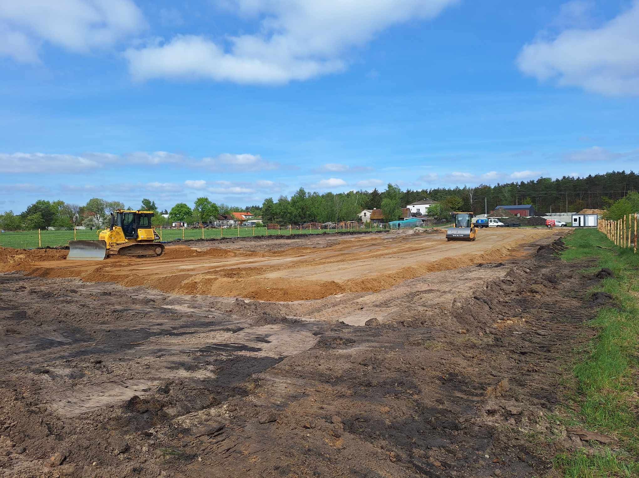 Żółty spychacz i walec na placu budowy, w tle zabudowania i drzewa, błękitne niebo z chmurami. Teren wyrównany, przygotowany pod dalsze prace.