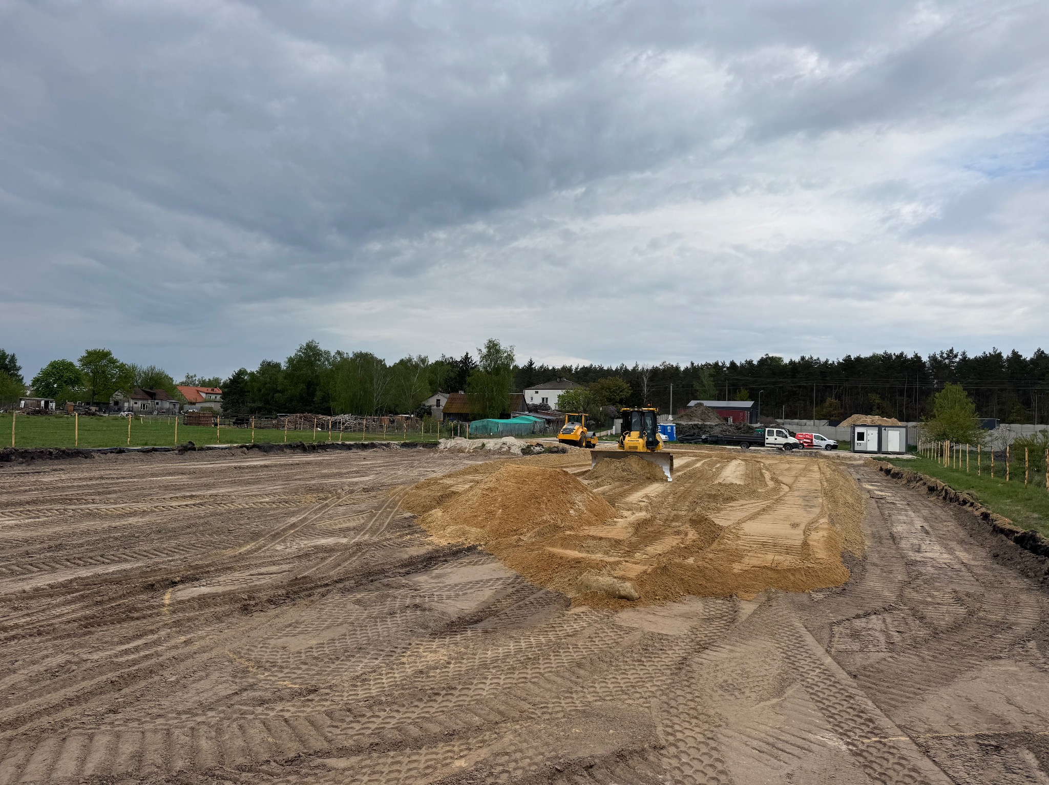 Żółta spycharka i walec na wyrównanym terenie z piaskiem, w tle zabudowania i pochmurne niebo. Widoczne ślady opon na ziemi.