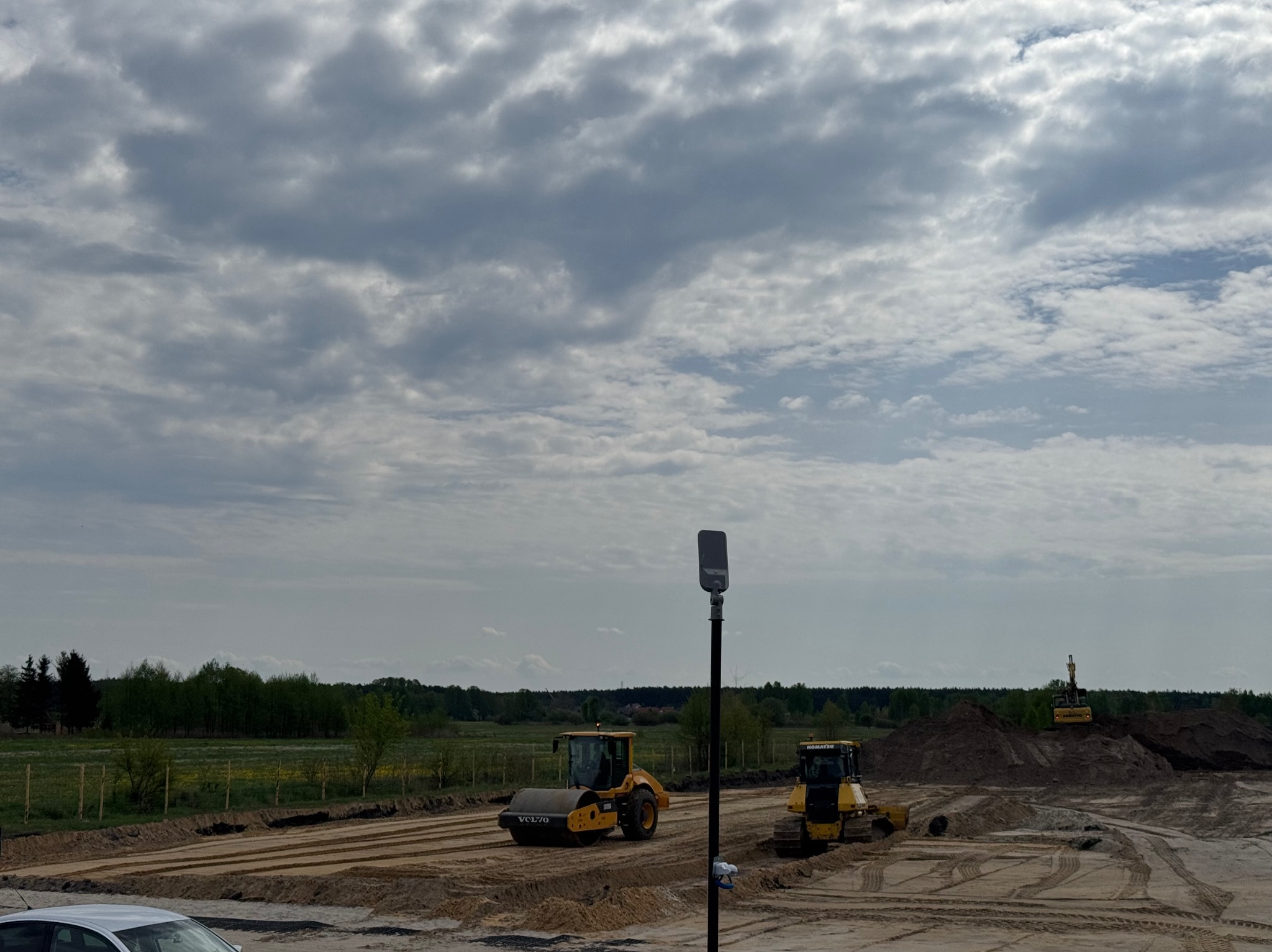 Żółty walec VOLVO i spychacz KOMATSU na placu budowy z ubitą ziemią i widoczną linią drzew w tle. Na wzgórzu koparka. Pochmurne niebo.