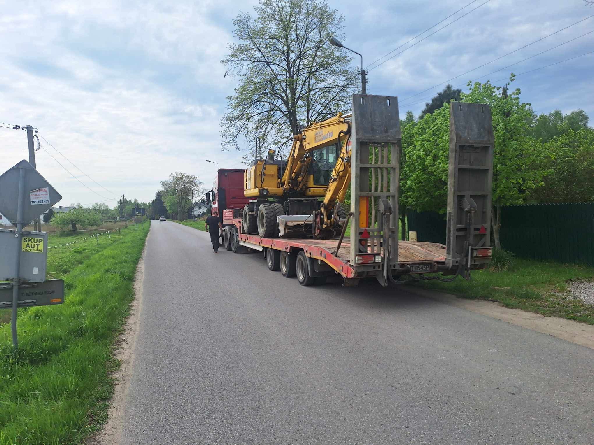 Transport niskopodwoziowy: żółta koparka kołowa na czerwonej platformie transportowej, zabezpieczona. Widok z tyłu na drogę z zielenią po bokach.
