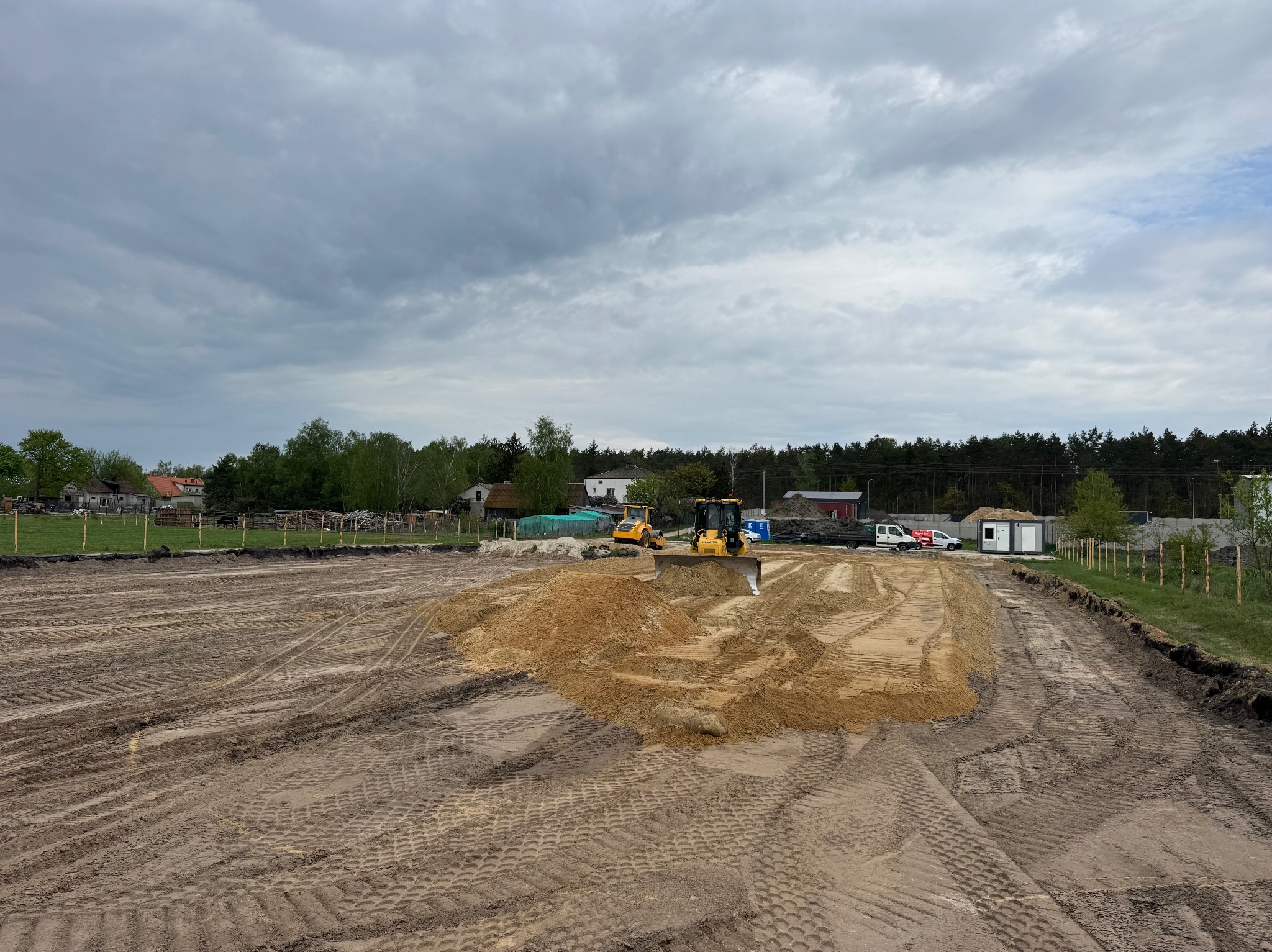 Żółta spycharka wyrównuje teren z piaskiem na budowie, widoczne ślady opon na ziemi. W tle budynki i zieleń. Pochmurne niebo.