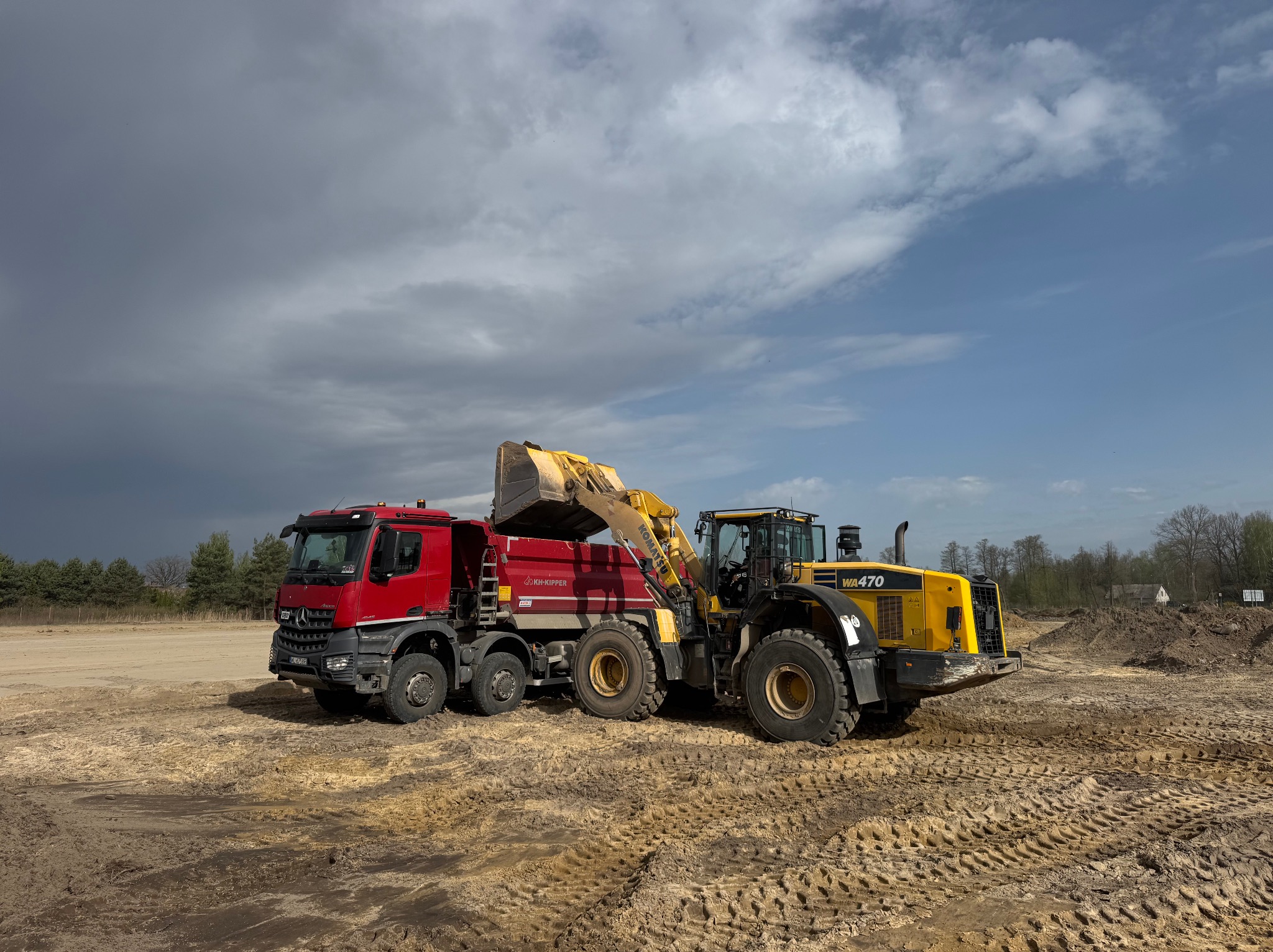 Żółta ładowarka Komatsu WA470 nabiera piasek do czerwonej wywrotki KH-Kipper na placu budowy pod pochmurnym niebem. Widoczne ślady opon na piasku.