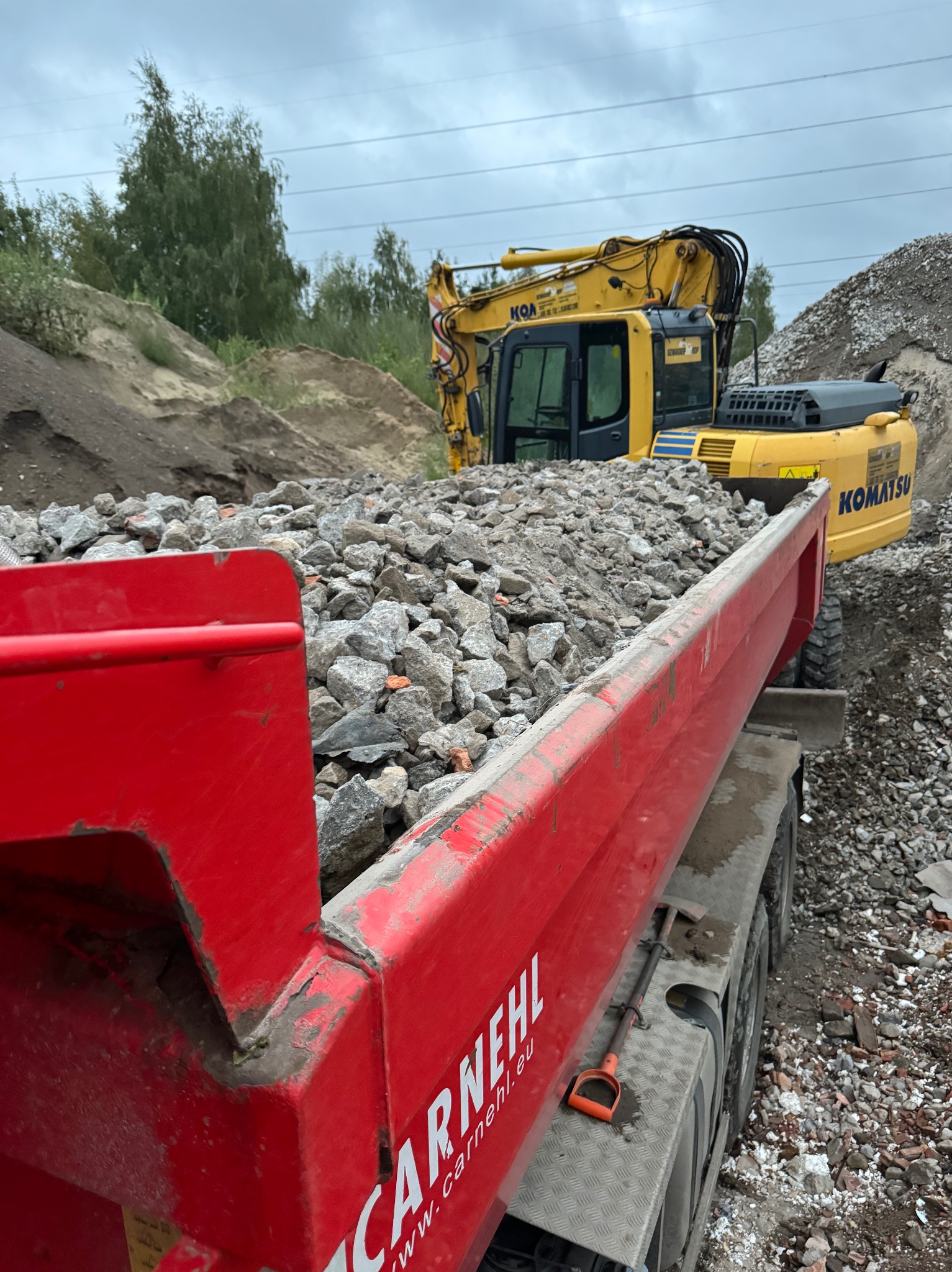 Czerwona ciężarówka wypełniona gruzem, z żółtą koparką Komatsu w tle, na terenie budowy z usypanymi hałdami ziemi i gruzu.