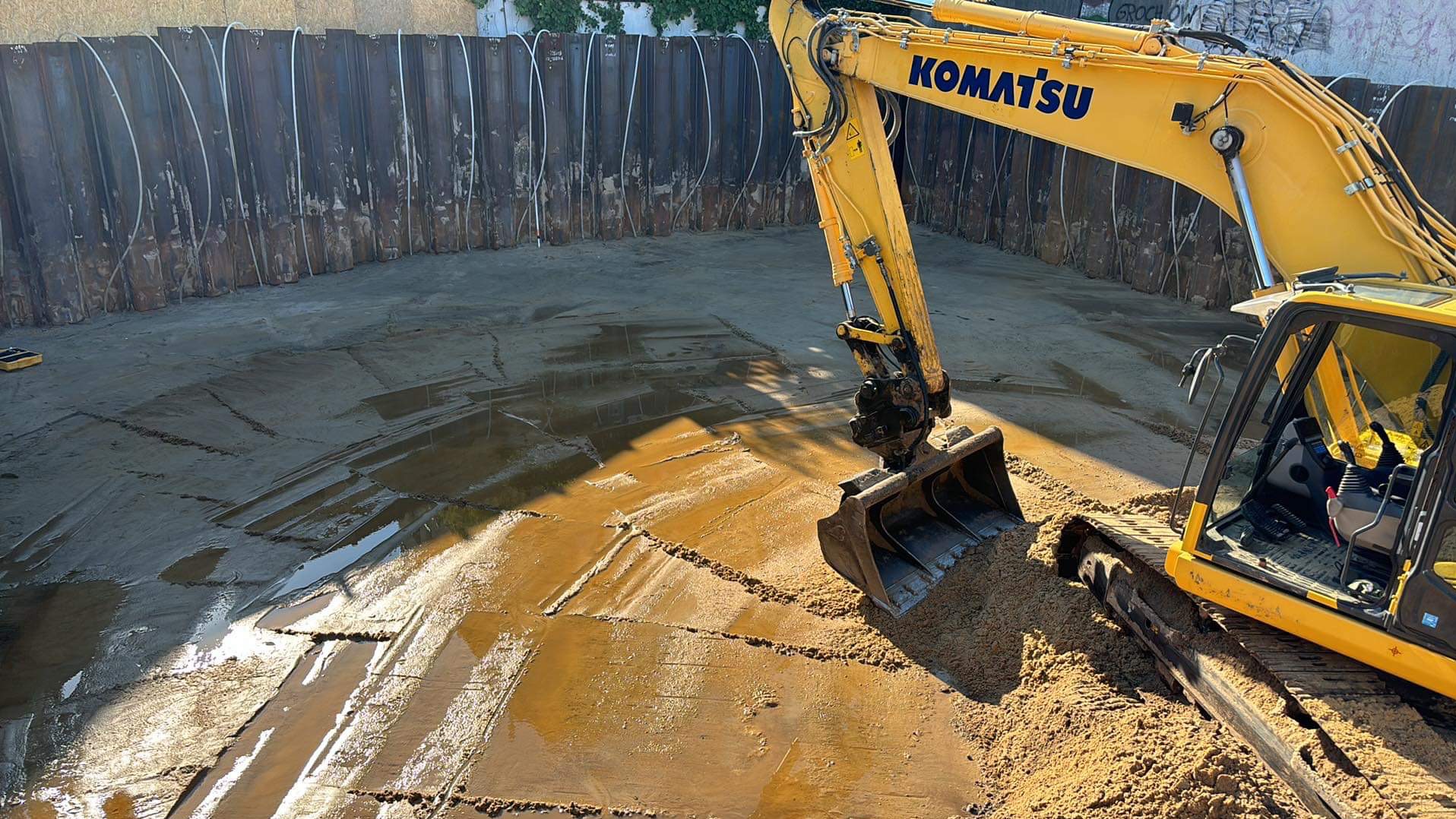 Żółta koparka Komatsu w wykopie zabezpieczonym ściankami Larsena. W tle widoczne białe rurki drenażowe. Ślady pracy łyżki koparki na mokrej ziemi.