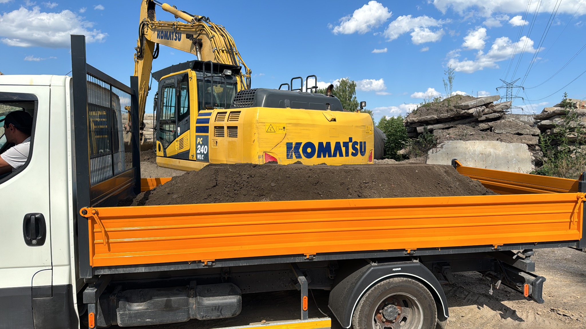 Żółta koparka Komatsu ładuje ziemię na pomarańczową ciężarówkę. W tle stos gruzu i błękitne niebo z chmurami.