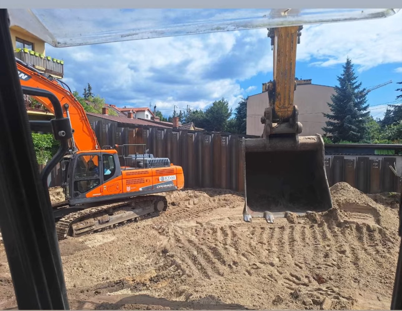 Pomarańczowa koparka Doosan DX340 pracuje w wykopie zabezpieczonym stalowymi ściankami Larsena. Widok z kabiny innej maszyny, w tle zabudowa jednorodzinna.