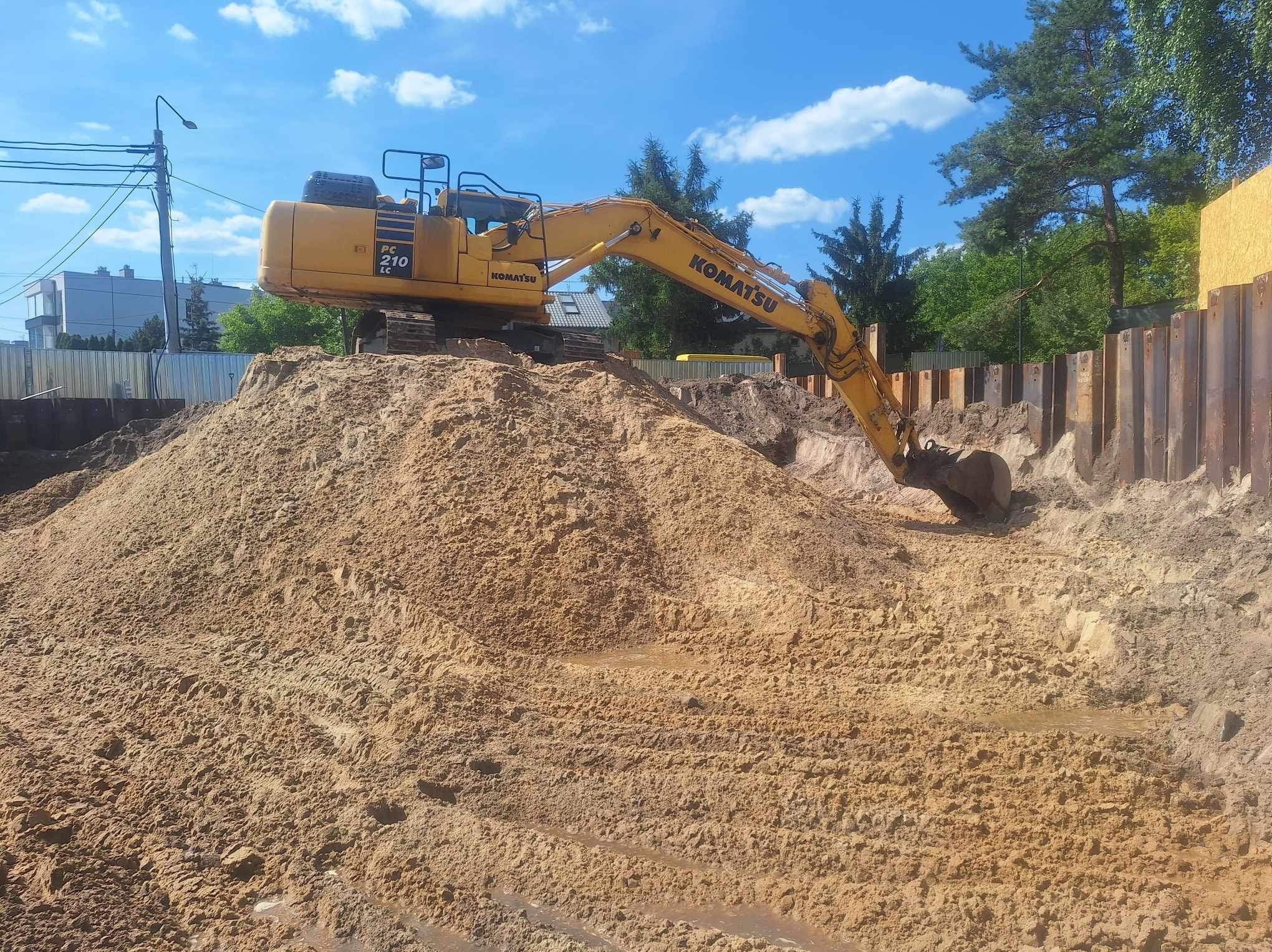 Żółta koparka Komatsu PC210LC nabiera piasek z hałdy. W tle widoczne stalowe ścianki wykopu, budynki i drzewa pod błękitnym niebem.