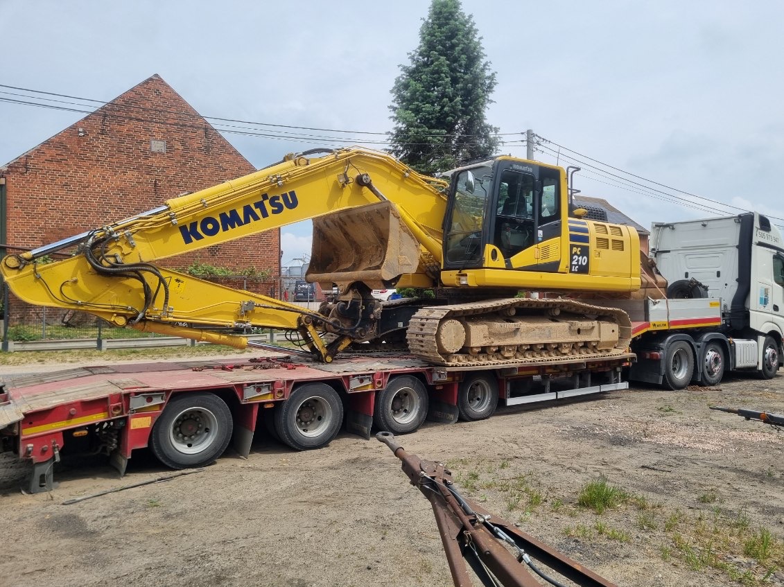 Żółta koparka Komatsu PC 210 LC, brudna od ziemi, transportowana na niskopodwoziowej przyczepie ciężarówki, z widocznym fragmentem haka holowniczego na pierwszym planie.