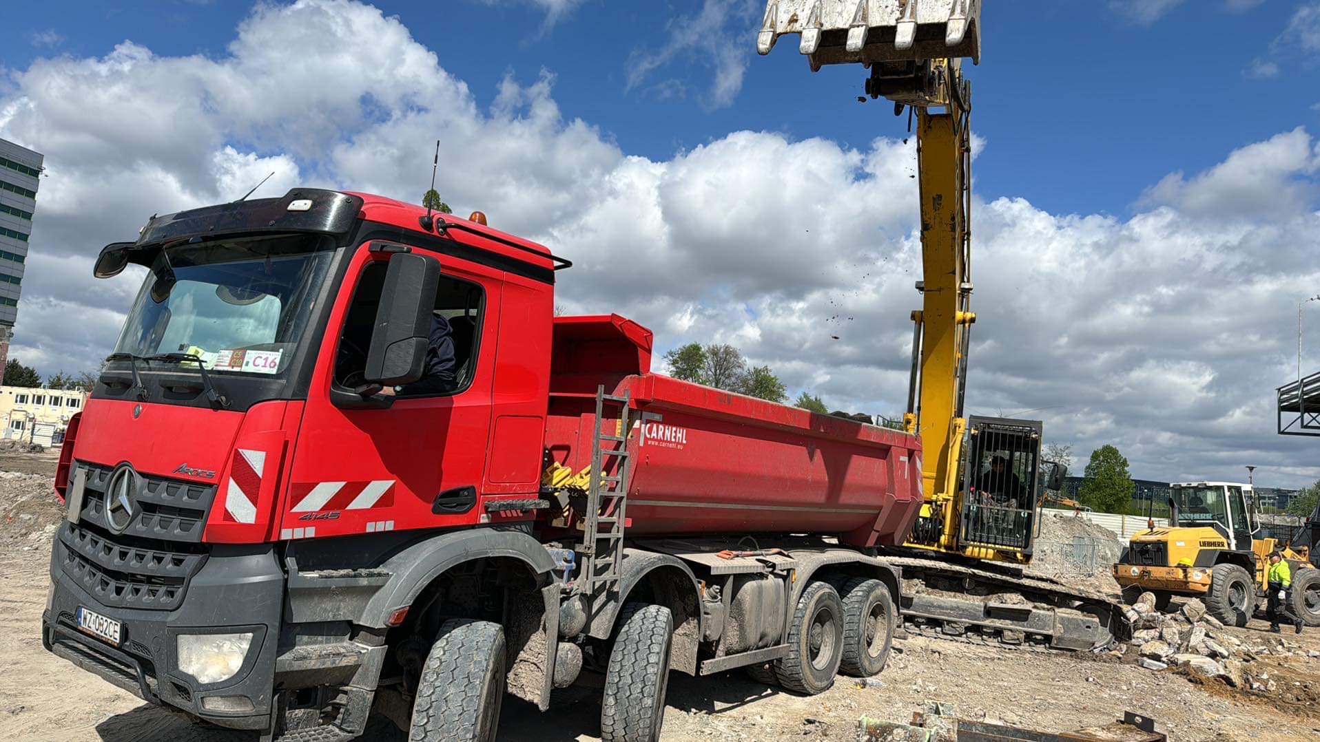 Czerwona ciężarówka i koparka w trakcie prac ziemnych na placu budowy, z widocznymi elementami infrastruktury i błękitnym niebem w tle.