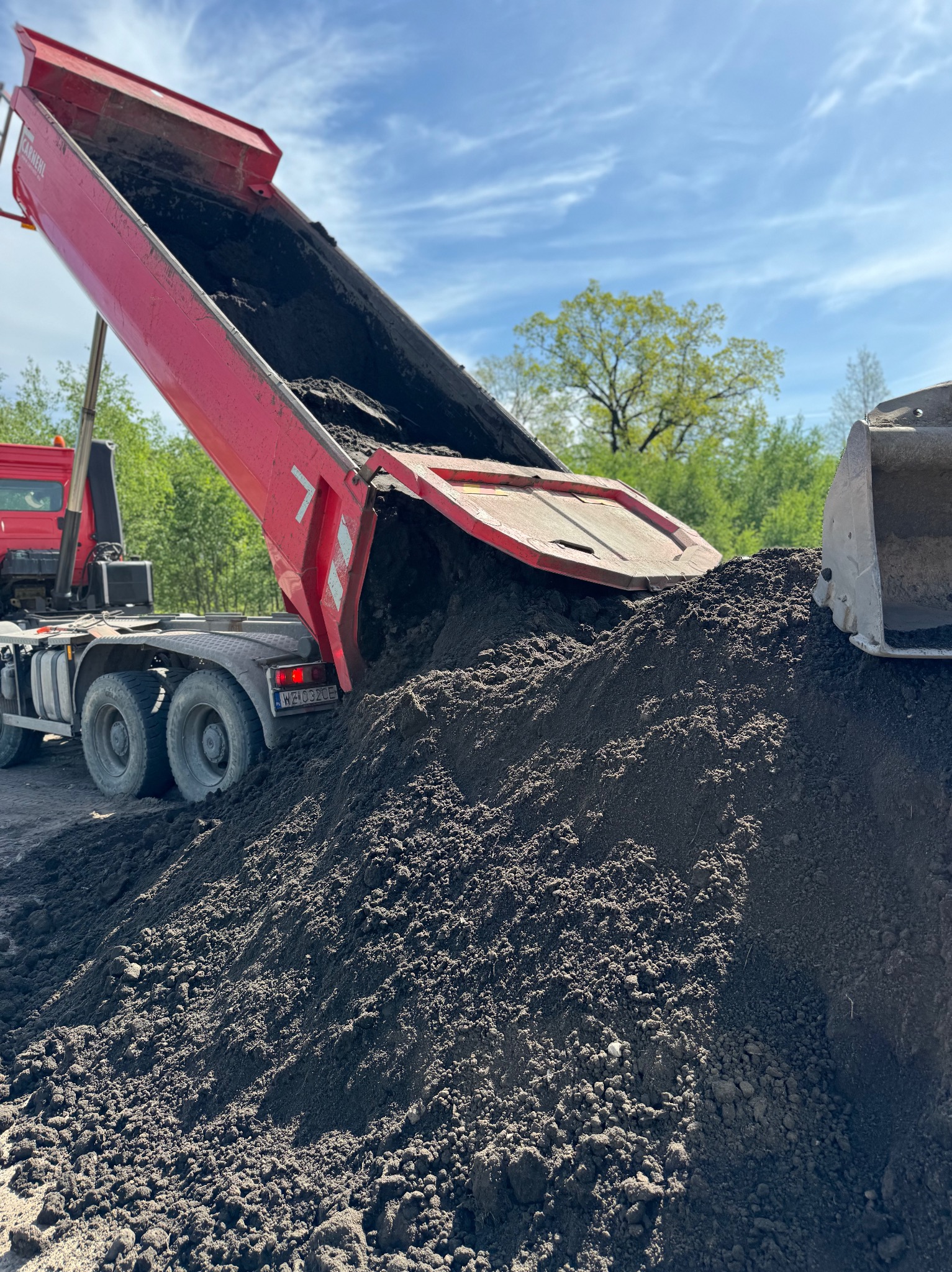 Czerwona wywrotka w trakcie wysypywania ciemnej ziemi, tworząc pokaźny nasyp na placu budowy, z zielenią drzew w tle i fragmentem koparki widocznym po prawej.