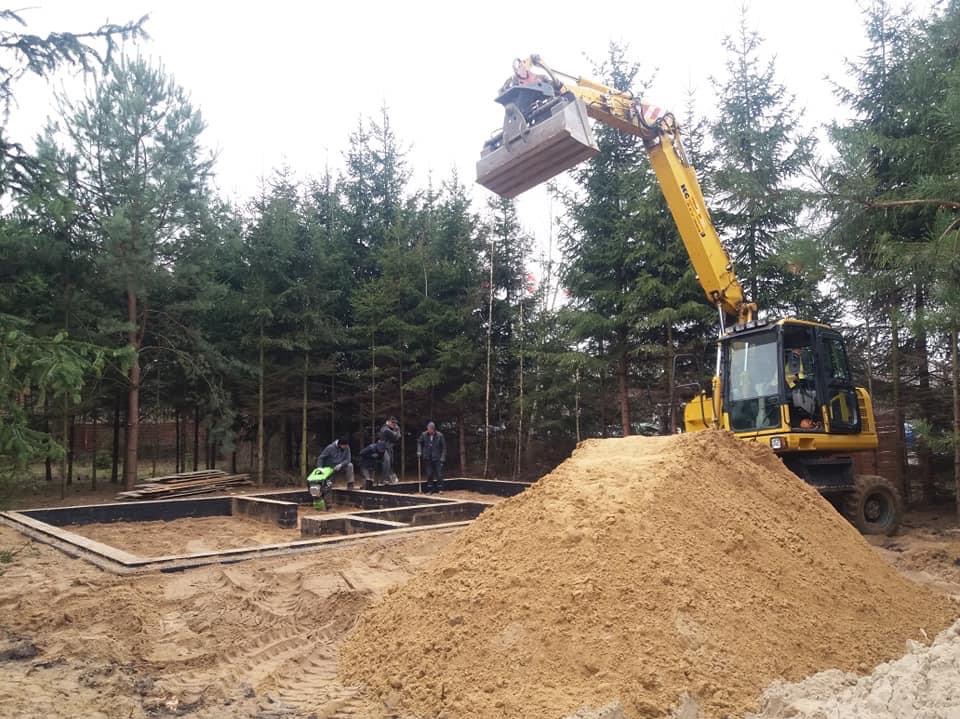 Żółta koparka z łyżką nad fundamentem domu, grupa robotników zagęszcza piasek w lesie. Stos piasku na pierwszym planie.