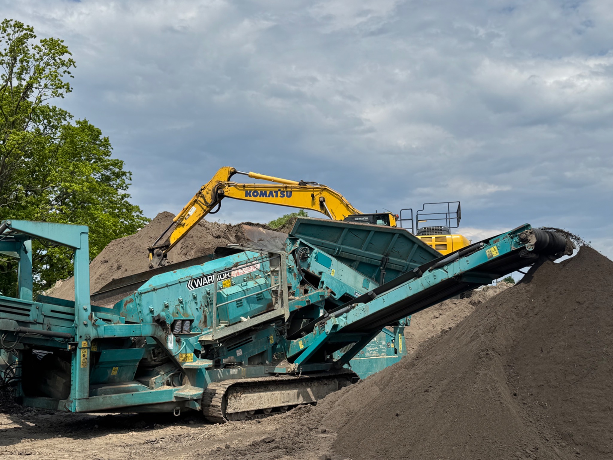 Praca przesiewacza mobilnego Warrior i koparki Komatsu na placu budowy. Urządzenia pracują na tle pochmurnego nieba i zieleni drzew.