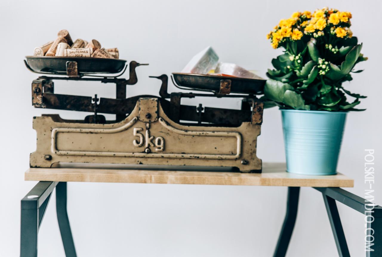 Antyczna waga szalkowa na drewnianym blacie, obciążona korkami od wina i zapakowanymi kostkami mydła, obok niebieska doniczka z kwitnącą na żółto rośliną.