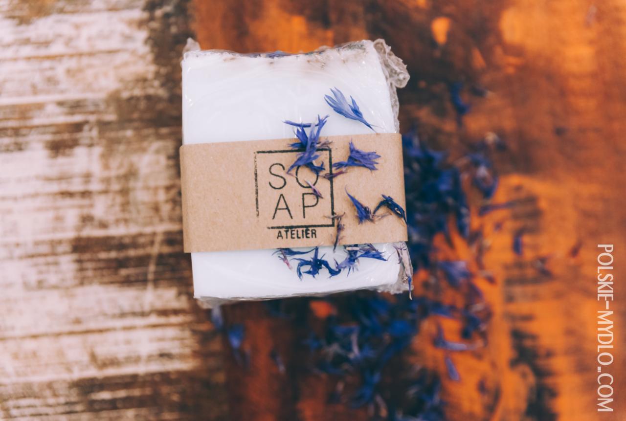 Ręcznie robione białe mydło owinięte papierową banderolą z napisem 'SOAP ATELIER', udekorowane suszonymi niebieskimi bławatkami, leżące na drewnianym tle z widocznym adresem strony internetowej.