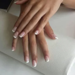 Dłonie z klasycznym, białym french na poduszce do manicure, obok leży smartfon.