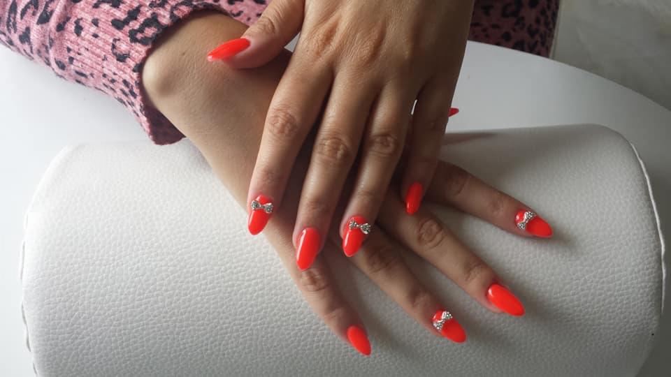 Dłonie z czerwonymi, szpiczastymi paznokciami ozdobionymi srebrnymi kokardkami na białym podłożu, widoczne z bliska.