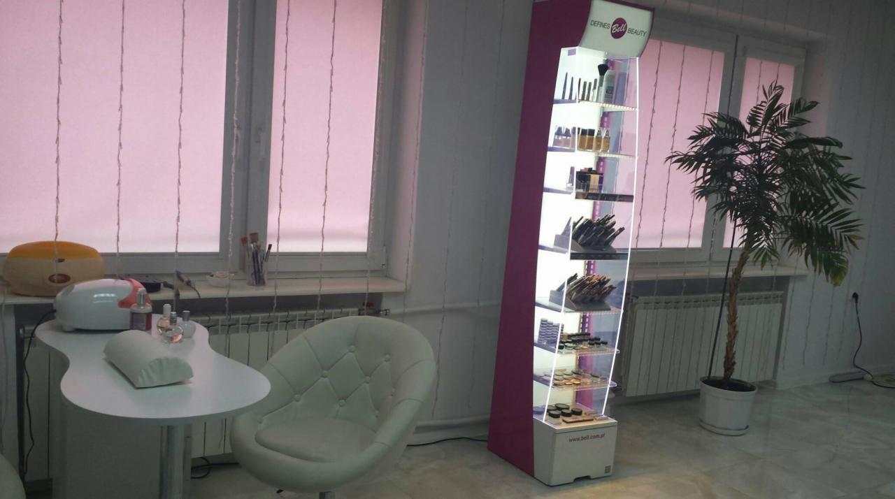 Stanowisko do stylizacji paznokci z lampą UV, poduszką pod dłoń i akcesoriami, obok regał z kosmetykami i palmą w doniczce, wnętrze salonu z oknami przysłoniętymi różowymi roletami.