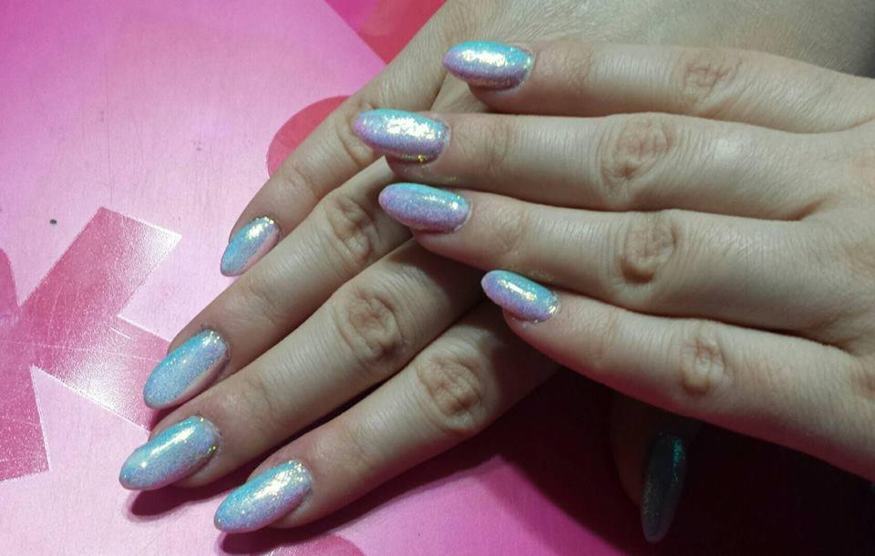 Dłonie z opalizującymi, błyszczącymi paznokciami w odcieniach błękitu i różu, ułożone na różowym tle.