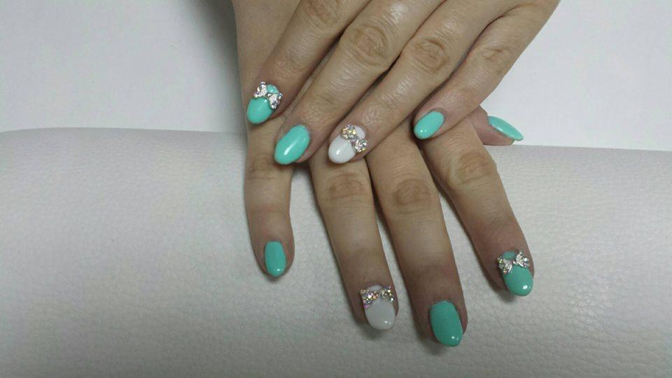 Dłonie z owalnymi paznokciami pomalowanymi na miętowy i biały kolor, dwa z nich ozdobione błyszczącymi kokardkami, ułożone na białym podłożu.