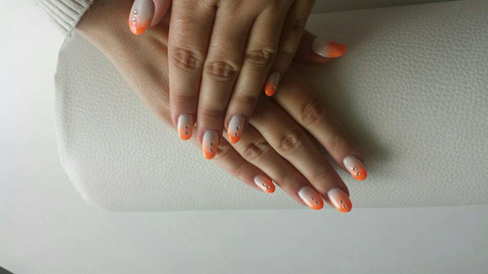 Dłonie z pomarańczowo-białym ombre na paznokciach, ozdobione małymi srebrnymi dżetami, ułożone na białym, skórzanym podłożu.