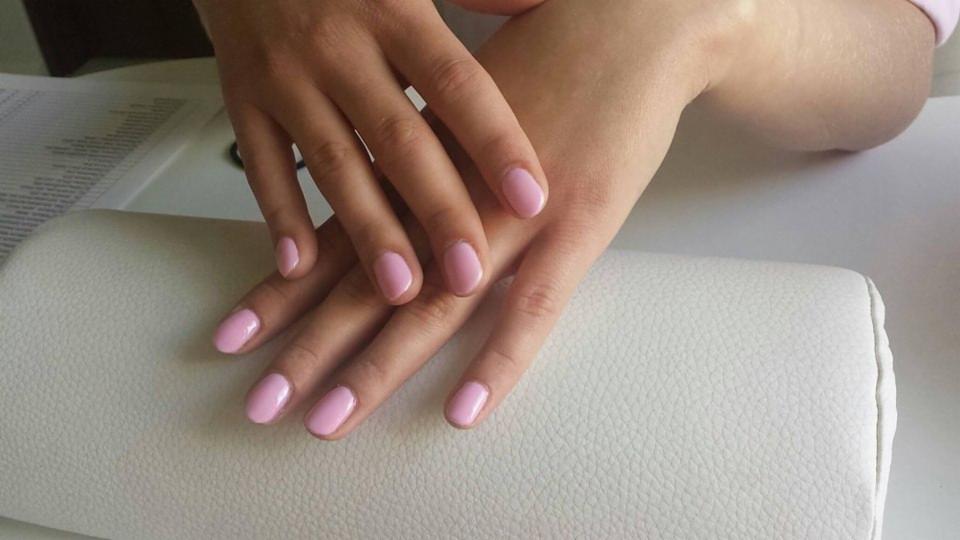 Dłonie z delikatnym, różowym lakierem hybrydowym na owalnych paznokciach, ułożone na białym podkładce pod ręce.