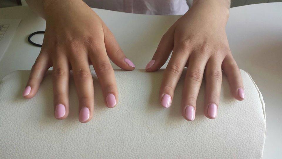 Dłonie z świeżo pomalowanymi na jasny róż paznokciami, ułożone na białym, skórzanym podłożu; w tle widoczna gumka do włosów i kartka.