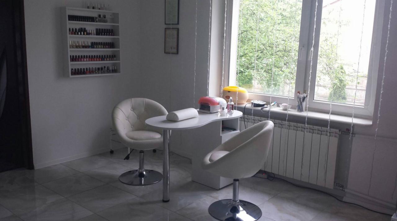 Stanowisko do stylizacji paznokci z białymi krzesłami, lampą UV i półką z lakierami w salonie kosmetycznym, widok z okna na zieleń.