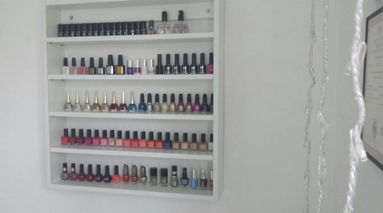 Półka wypełniona różnorodnymi lakierami do paznokci w odcieniach czerwieni, różu, złota, srebra i czerni, ustawionymi na białym tle ściany, z delikatnymi ozdobami w postaci girlandy po prawej stronie.