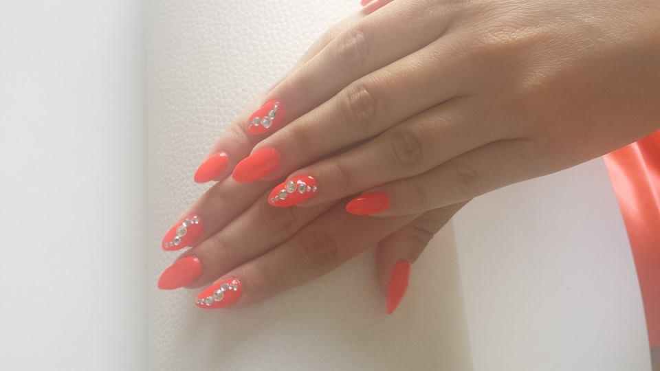 Pomarańczowe paznokcie w kształcie migdała zdobione rzędem srebrnych cyrkonii na tle białej skóry dłoni i fragmentu pomarańczowej tkaniny.