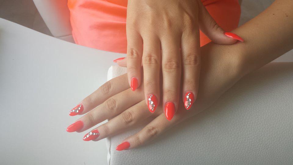 Dłonie z czerwonymi, migdałowymi paznokciami ozdobionymi rzędami błyszczących, srebrnych cyrkonii na tle białego fotela i neonowo-pomarańczowej bluzki.