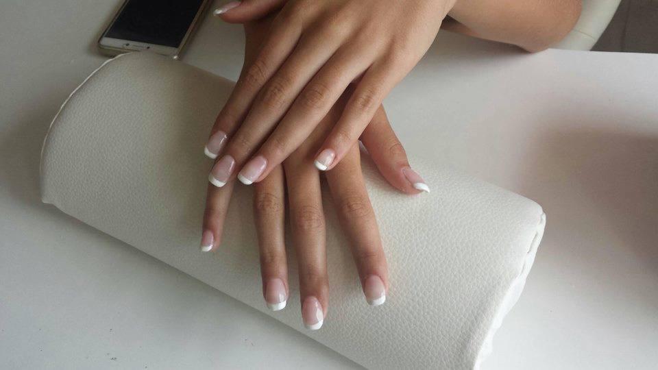 Dłonie z klasycznym, białym french na poduszce do manicure, obok leży smartfon.