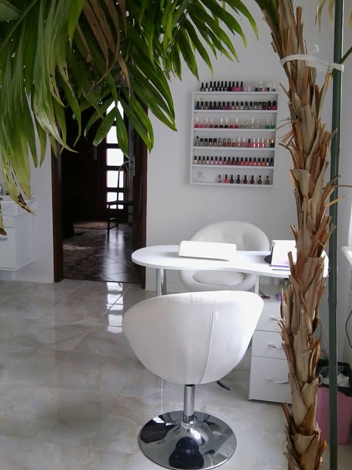 Jasne wnętrze salonu kosmetycznego z białym stołem do zabiegów, krzesłami z chromowaną podstawą, ścianą z półkami pełnymi lakierów do paznokci i dekoracyjną palmą.
