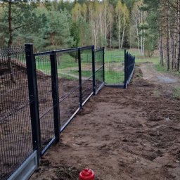 OGRO-RAD - Czarny system ogrodzeniowy z siatki z betonową podmurówką, widok na las w tle, czerwony hydrant na pierwszym planie.