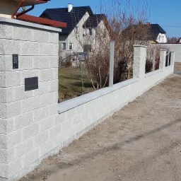 Nowoczesne ogrodzenie z szarego betonu modułowego z wbudowaną skrzynką na listy i panelem domofonowym, widok z zewnątrz na tle domów jednorodzinnych.