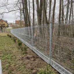 Długi, szary system ogrodzeniowy z siatki, z betonową podmur&oacute;wką, ciągnący się wzdłuż działki w Ostrowi Mazowieckiej, z młodymi drzewkami posadzonymi wzdłuż linii ogrodzenia.