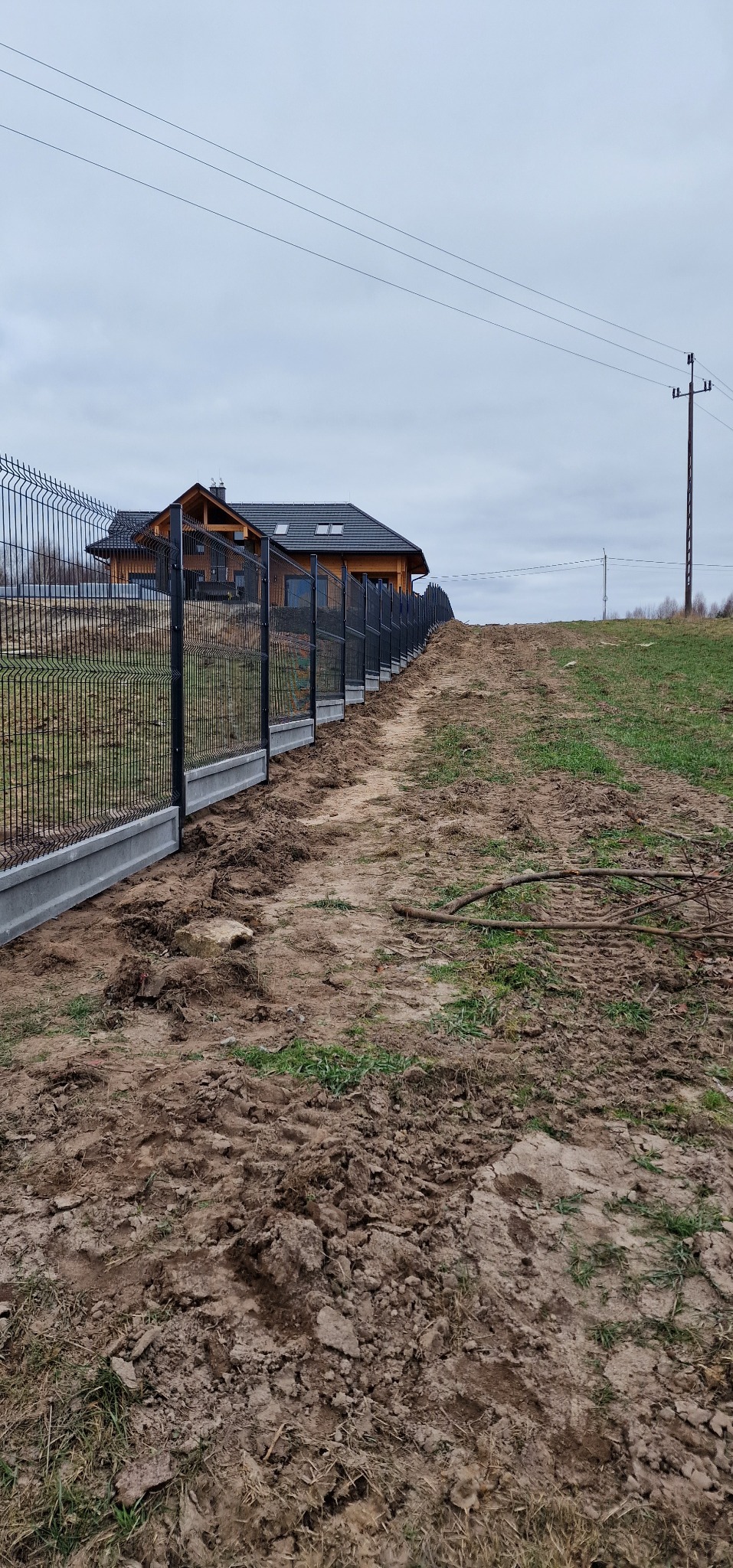 Długi, czarny ogrodzenie panelowe z betonową podmurówką, prowadzące wzdłuż polnej drogi w kierunku drewnianego domu pod pochmurnym niebem.