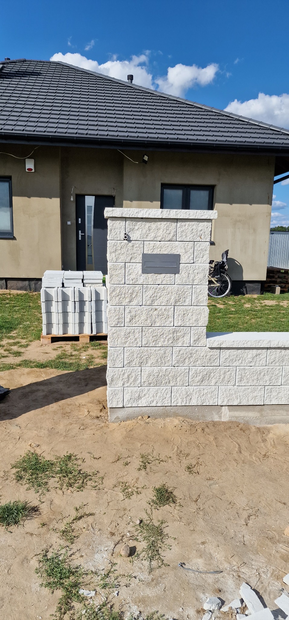 Fragment murku z jasnych bloczków betonowych z zamontowaną skrzynką na listy w Ostrowi Mazowieckiej, obok paleta z bloczkami, w tle dom jednorodzinny.