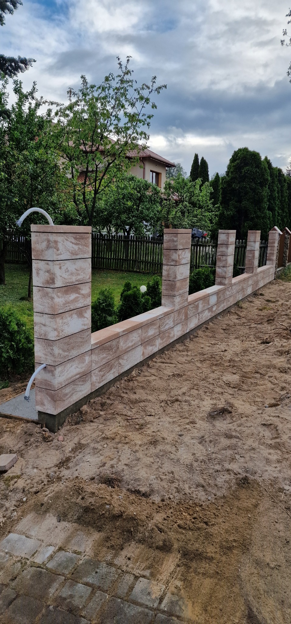 Wznoszona konstrukcja ogrodzenia z bloków betonowych o teksturze piaskowca, widoczny biały przewód wystający z pierwszego słupka, w tle drewniane ogrodzenie posesji i zabudowa jednorodzinna