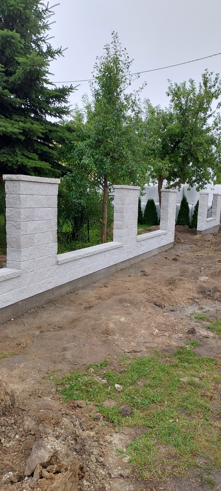 Niski, modułowy mur z szarego, betonowego bloczka o nieregularnej fakturze, oddzielający posesję od drogi gruntowej, z młodymi drzewami w tle.