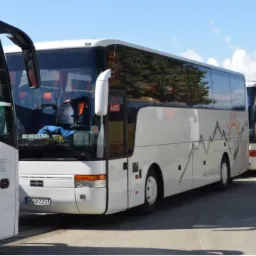 Biały autokar z grafiką gór i słońca na boku, zaparkowany obok innych autokarów, widoczny numer rejestracyjny.