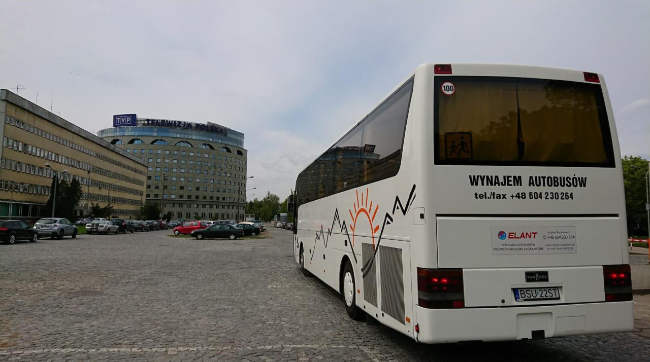 Biały autokar z grafiką gór i słońca, zaparkowany na brukowanym placu z budynkami w tle, widoczny numer kontaktowy 'Wynajem Autokarusów'.