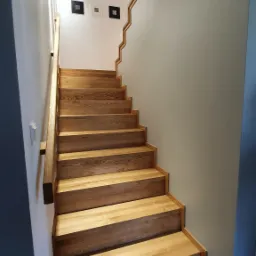 Jasne, drewniane schody z poręczą, oświetlone punktowymi lampami na suficie, prowadzące do góry. Na ścianie widoczne trzy małe, kwadratowe obrazy w czarnych ramkach.