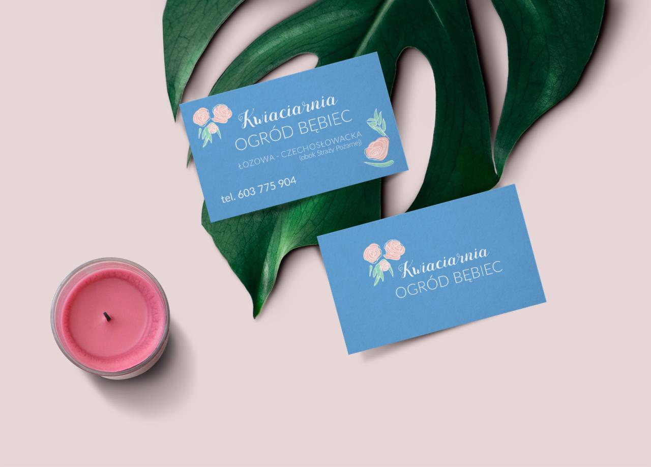 Dwie błękitne wizytówki kwiaciarni Ogród Bębiec, z motywem róż i liści, ułożone na tle zielonego liścia monstery i różowej świecy.
