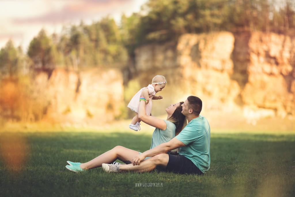 Szczęśliwa rodzina siedząca na trawie: mama podnosi w górę uśmiechniętą córeczkę, tata obejmuje mamę, w tle skalny klif i drzewa.