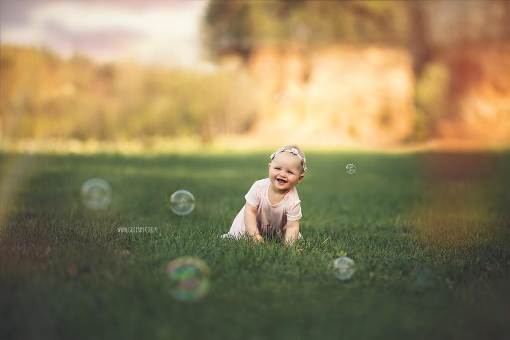 Uśmiechnięta niemowlęca dziewczynka z wianuszkiem na głowie raczkuje po trawie, otoczona bańkami mydlanymi na tle rozmytego pleneru z drzewami.