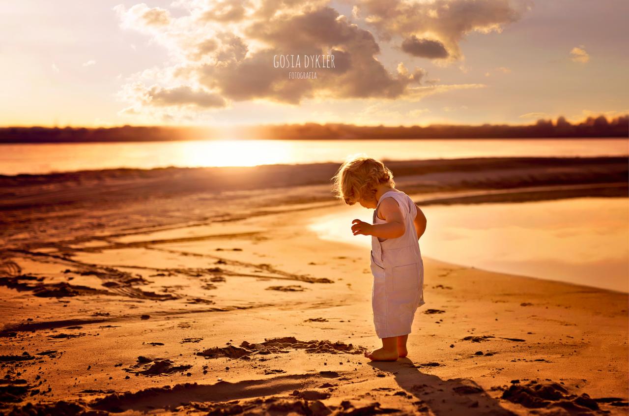 Mała dziewczynka w jasnym kombinezonie bawi się na piaszczystej plaży o zachodzie słońca, obserwując coś w piasku.