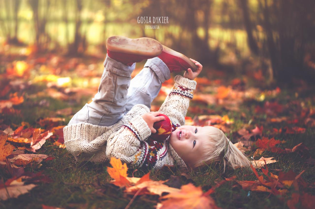 Mała dziewczynka w grubym swetrze leży na plecach wśród jesiennych liści, trzymając jabłko i podnosząc nogi w czerwonych butach do góry.