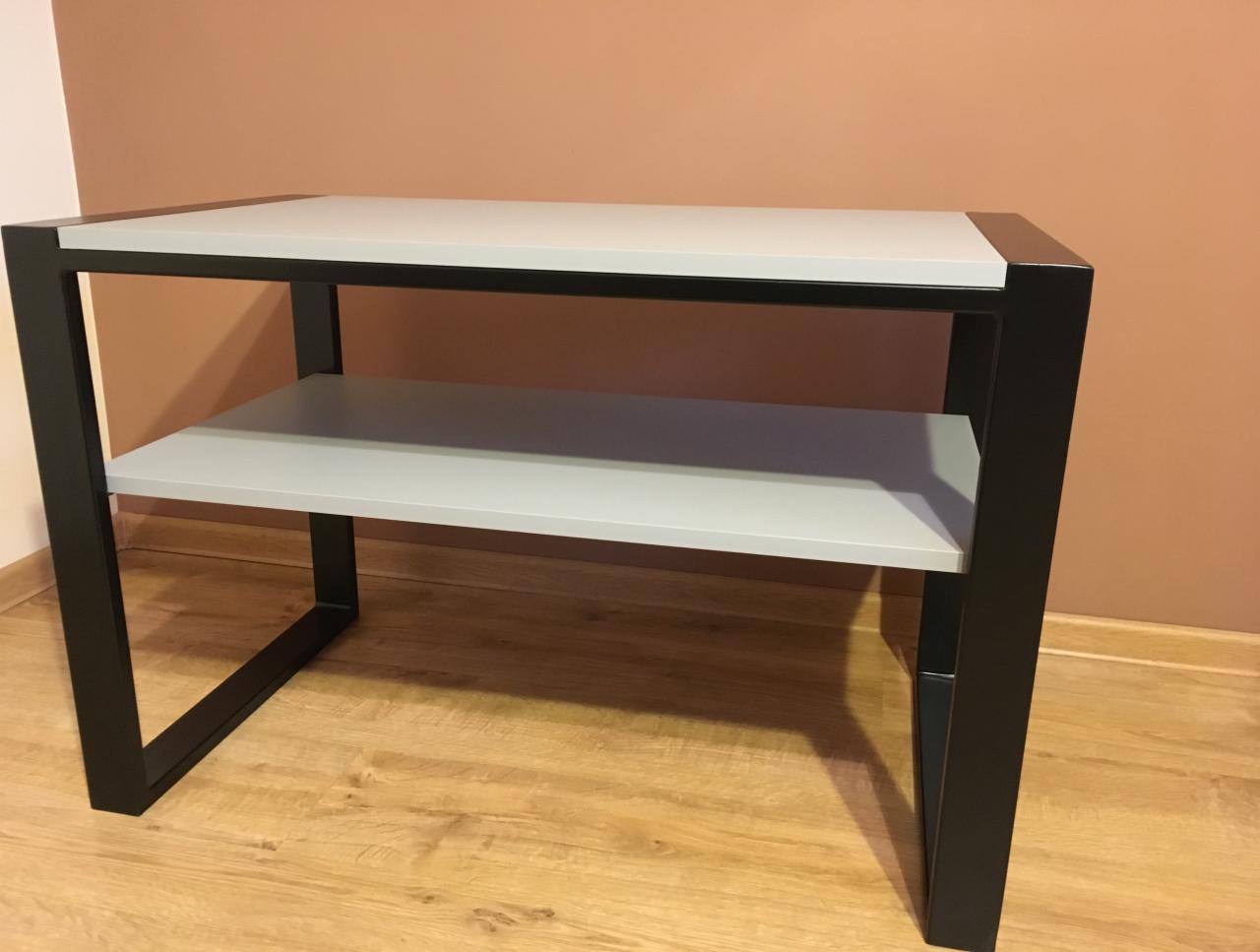 Dwupoziomowy stolik RTV na czarnej metalowej ramie z blatem i półką w odcieniach szarości, na drewnianej podłodze i tle ściany w kolorze terakoty.