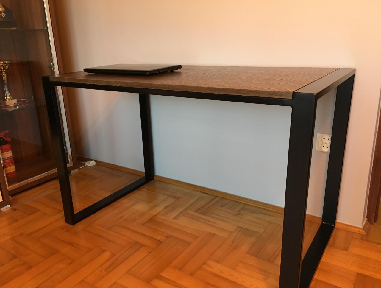Biurko z blatem drewnianym i czarnymi, metalowymi nogami w stylu loft, na blacie zamknięty laptop, parkiet w jodełkę na podłodze.