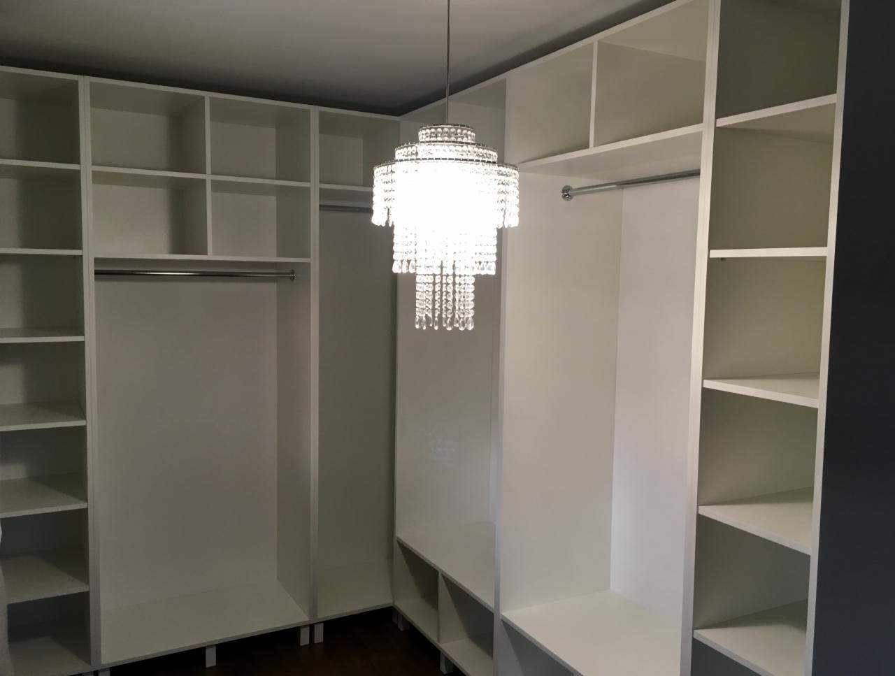 Biała garderoba z półkami i drążkami, oświetlona kryształowym żyrandolem, widok z dołu, minimalistyczny design.